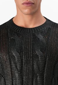 Nærbilde av en person som har på seg en svart, åpenstrikket genser med store flettede kabelmønstre og ribbestrikket rund hals.