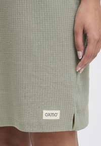 Kéz egy világoszöld, texturált szoknya mellett, amely kis oldalsó hasítékkal rendelkezik, és az alján az "oxmo" címke található.