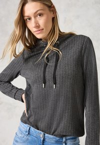 Pullover gris foncé avec des rayures verticales, en tissu léger, capuche avec cordons de serrage, et manches longues, associé à un jean bleu clair.