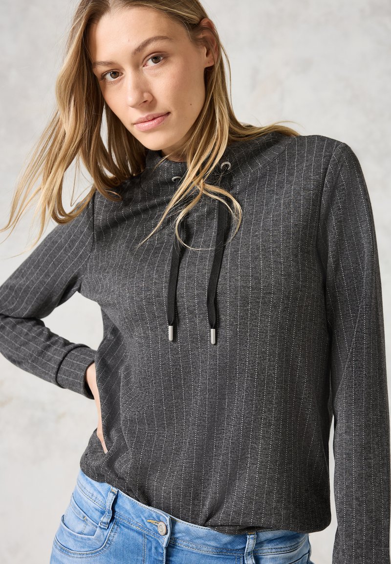 Pullover gris foncé avec des rayures verticales, en tissu léger, capuche avec cordons de serrage, et manches longues, associé à un jean bleu clair.