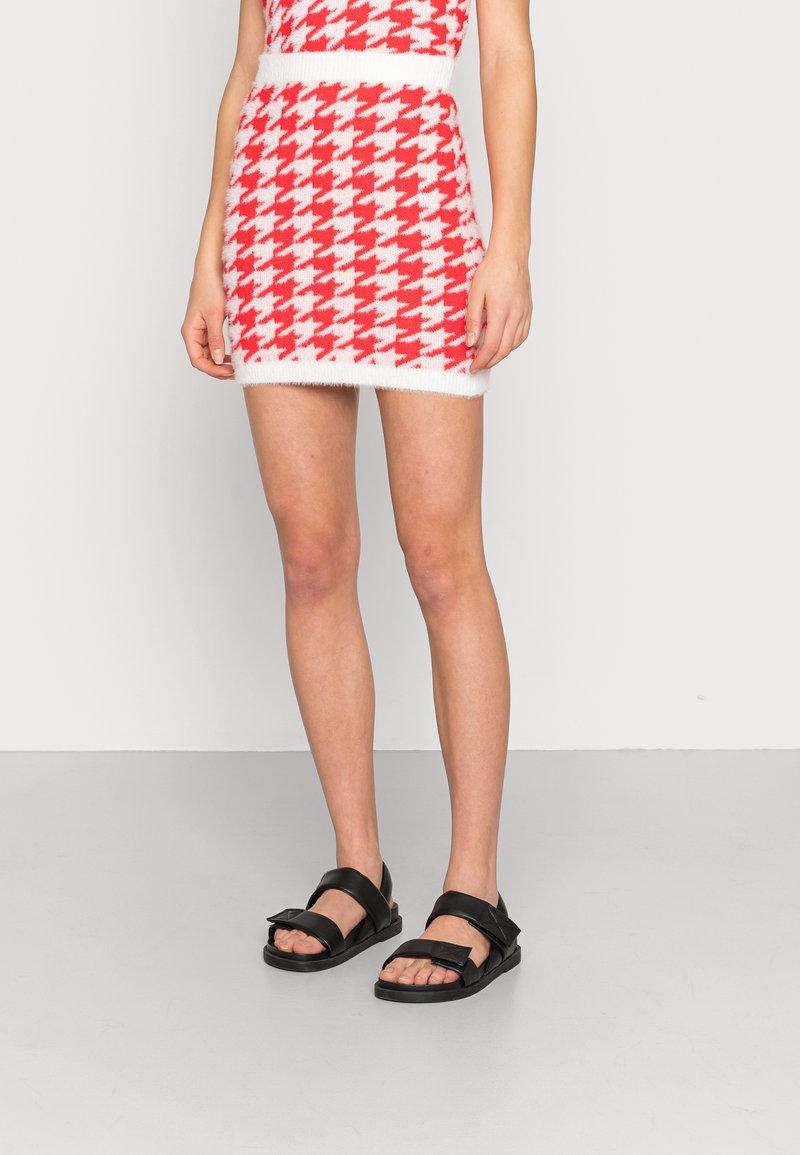 Mini saia com padrão pied-de-poule em vermelho e branco, feita de material de malha suave, com uma cintura branca e combinada com sandálias pretas.