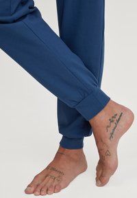 Middagblauwe, lichtgewicht broeken met elastische boorden weergegeven naast een blote voet met meerdere tatoeages. Glad textuur van de stof is zichtbaar.