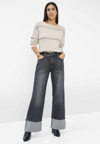 Maglione a righe crema e grigio, jeans a gamba larga di un colore scuro con risvolti in denim chiaro, stivali neri con tacco, modella in piedi con le mani in tasca.
