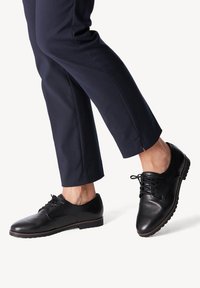 Chaussures noires en cuir à lacets avec orteils ronds et semelles texturées, portées avec un pantalon bleu marine présentant une légère fente à l'ourlet.