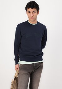 Felpa navy con logo "BELSTAFF" in un blu navy più scuro, scollatura tonda. Indossata sopra una camicia verde chiaro e abbinata a jeans grigi.