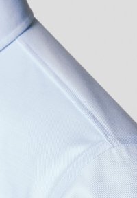 Tkanina w jasnoniebieskim kolorze z teksturowanym, żebrowanym wzorem. Zbliżenie pokazuje detale szwów oraz gładkie wykończenie kołnierza i ramienia.