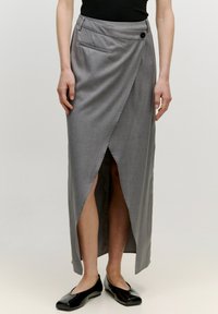 Grauer Wickelrock aus glattem Material, mit einem seitlichen Schlitz, Taschen-Detail und einem Knopfverschluss an der Taille. Tragen mit schwarzen Ballerinas.