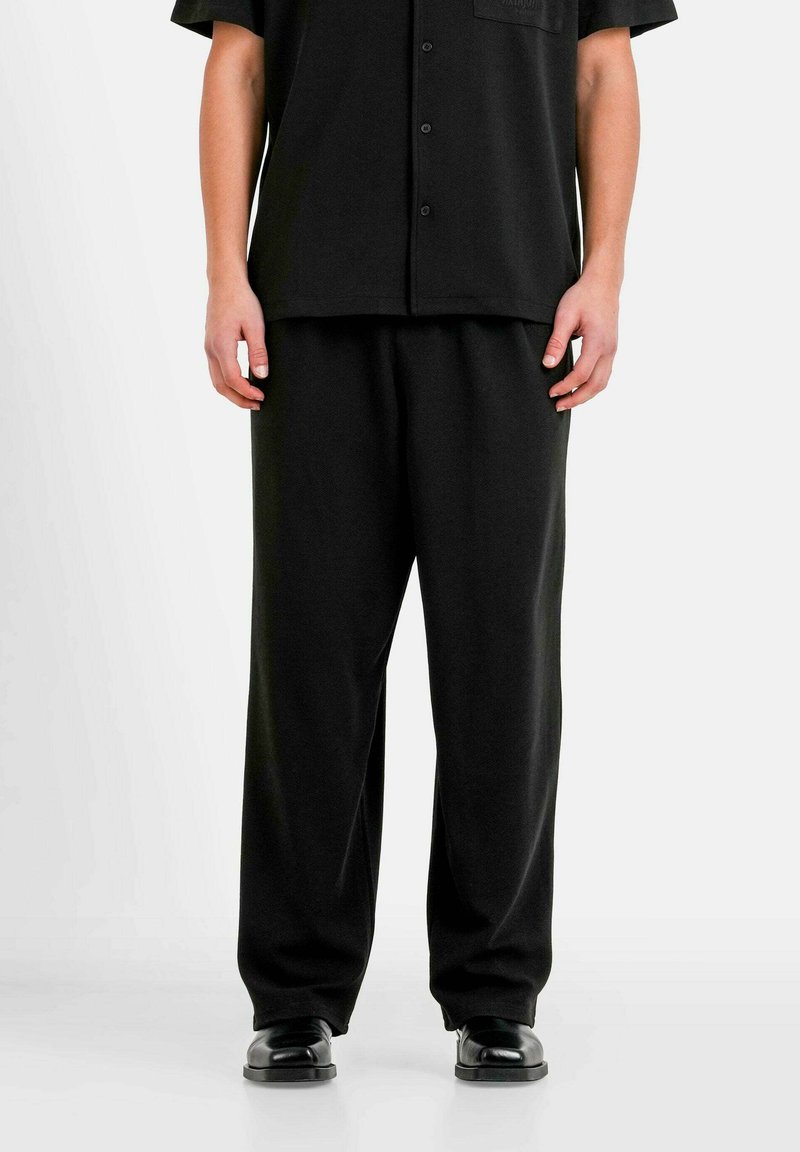 Chemise noire à manches courtes avec un design à boutons et une poche poitrine, associée à un pantalon noir ample et des chaussures noires brillantes.