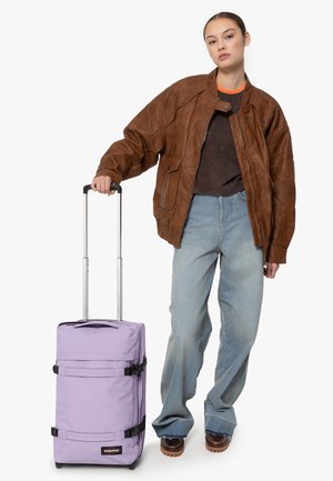Purple rolling suitcase with black straps, sturdy handle, and compact design. Model wearing a brown leather jacket and loose jeans.