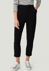 Pantalons noirs fuselés avec une texture lisse, dotés d'un devant plat, de passants de ceinture et de poches latérales. Associés à des baskets argentées métalliques.