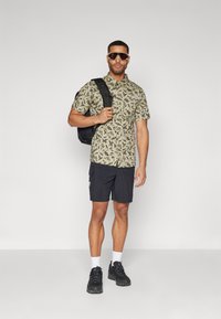 Korte mouwen, groen patroon shirt gecombineerd met zwarte cargoshorts. Een zwarte rugzak en zonnebril maken de outfit compleet. Sneakers met witte sokken.