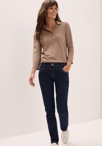 Mujer sonriendo, caminando, vestida con una camisa de polo de manga larga beige, jeans azules oscuros y zapatillas blancas, con una mano en el bolsillo.