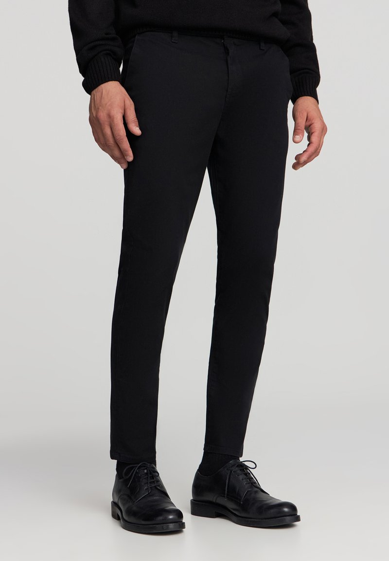 Czarne dopasowane spodnie o klasycznym kroju, z paskami na pasek i zamkiem błyskawicznym, w połączeniu z czarnymi skórzanymi butami. Gładka tekstura materiału.