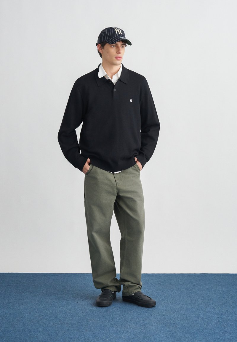 Junger Mann, der ein schwarzes Langarm-Poloshirt, olivgrüne Hose, schwarze Schuhe und eine schwarz gestreifte Baseballkappe trägt, steht mit den Händen in den Taschen.