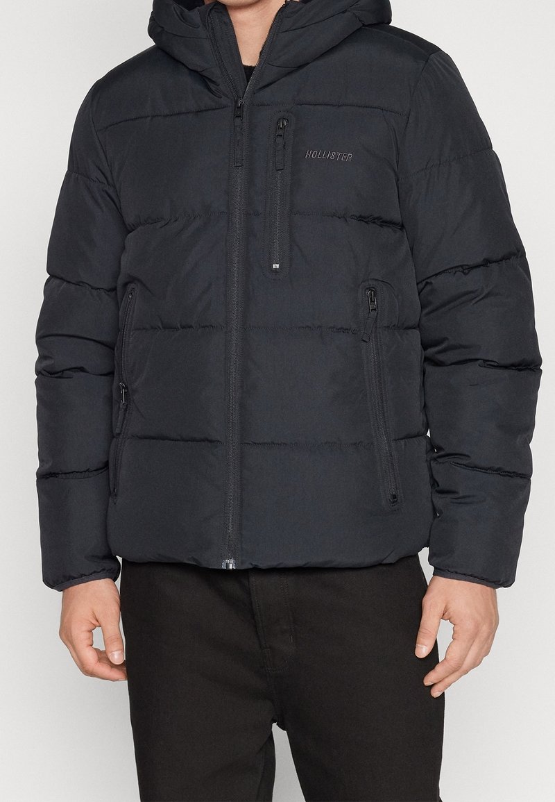 Schwarze Steppjacke mit Kapuze, mit horizontalen Nähten, zwei Seitentaschen mit Reißverschluss und einer Reißverschlusstasche auf der Brust. Der Stoff wirkt glatt.