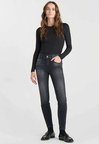 Haut noir à manches longues associé à un jean gris foncé à coupe slim. Le jean comporte des poches avant et une fermeture par bouton. Le modèle porte des mocassins noirs.
