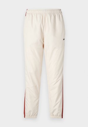 Pantalones de chándal color beige claro con cintura elástica, que presentan franjas rojas a los lados y un pequeño logo. Textura suave y brillante. Tobillos recogidos.