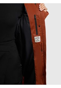 Chaqueta exterior marrón con interior negro. Cuenta con un bolsillo con cremallera y una etiqueta de tela. El material parece liso y resistente al agua.