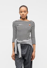 Νεαρή γυναίκα με πλεκτά μαλλιά που φοράει ριγέ μαύρο και λευκό πουκάμισο με μια μικρή κόκκινη καρδιά, γκρι πουλόβερ δεμένο στη μέση και σκουρόχρωμο παντελόνι.