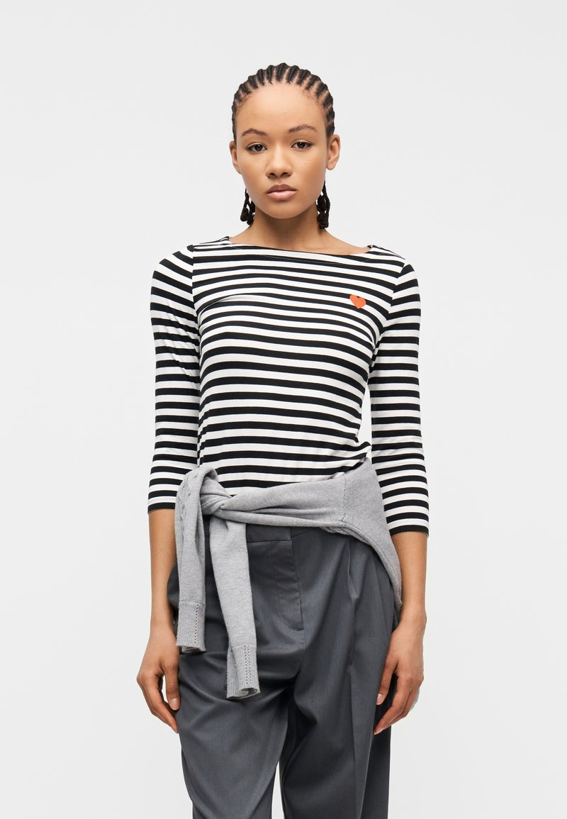 Νεαρή γυναίκα με πλεκτά μαλλιά που φοράει ριγέ μαύρο και λευκό πουκάμισο με μια μικρή κόκκινη καρδιά, γκρι πουλόβερ δεμένο στη μέση και σκουρόχρωμο παντελόνι.