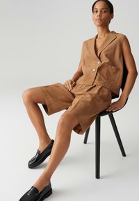 Braunes Leinen-Top ohne Ärmel mit zwei Knöpfen und einer Brusttasche, kombiniert mit passender Shorts. Das Model trägt schwarze Slipper.