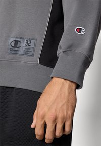 Sudadera gris con acentos negros, puños acanalados y un parche rectangular del logo de Champion que incluye texto. Una mano reposa sobre una tela negra texturada.
