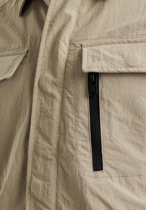 Veste beige en tissu texturé, dotée d'une grande poche à rabat et d'une poche latérale zippée avec un zip noir. Design simple et fonctionnel.
