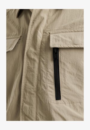 Beige Jacke aus strukturiertem Stoff, mit großer Klappentasche und einer seitlichen Tasche mit Reißverschluss und schwarzem Zipper. Einfaches, funktionales Design.