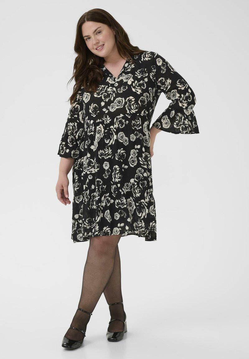 Schwarzes Blumenkleid mit weißem Rosendruck, V-Ausschnitt, dreiviertellange Glockenärmel und ausgestelltem Saum; kombiniert mit schwarzen Strumpfhosen und Absatzschuhen.