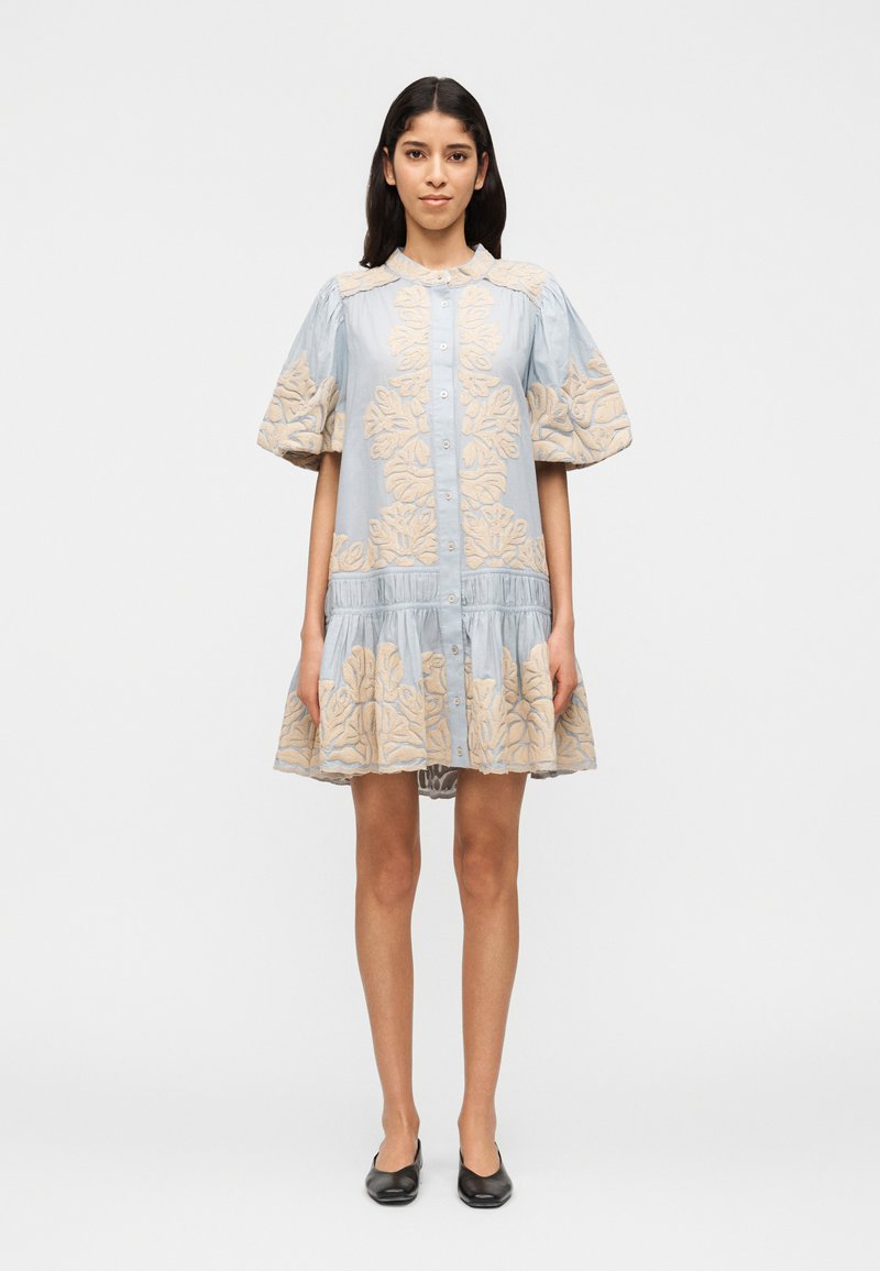 Γυναίκα που στέκεται, φορώντας ένα ανοιχτό μπλε  φόρεμα με κουμπιά και φουσκωτά μανίκια, με μπεζ λουλουδένια κεντήματα, συνδυασμένο με μαύρα παπούτσια φλατ.