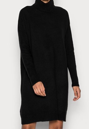 Femme portant une longue robe-pull noire à col roulé avec des manches longues, debout devant un fond clair uni.