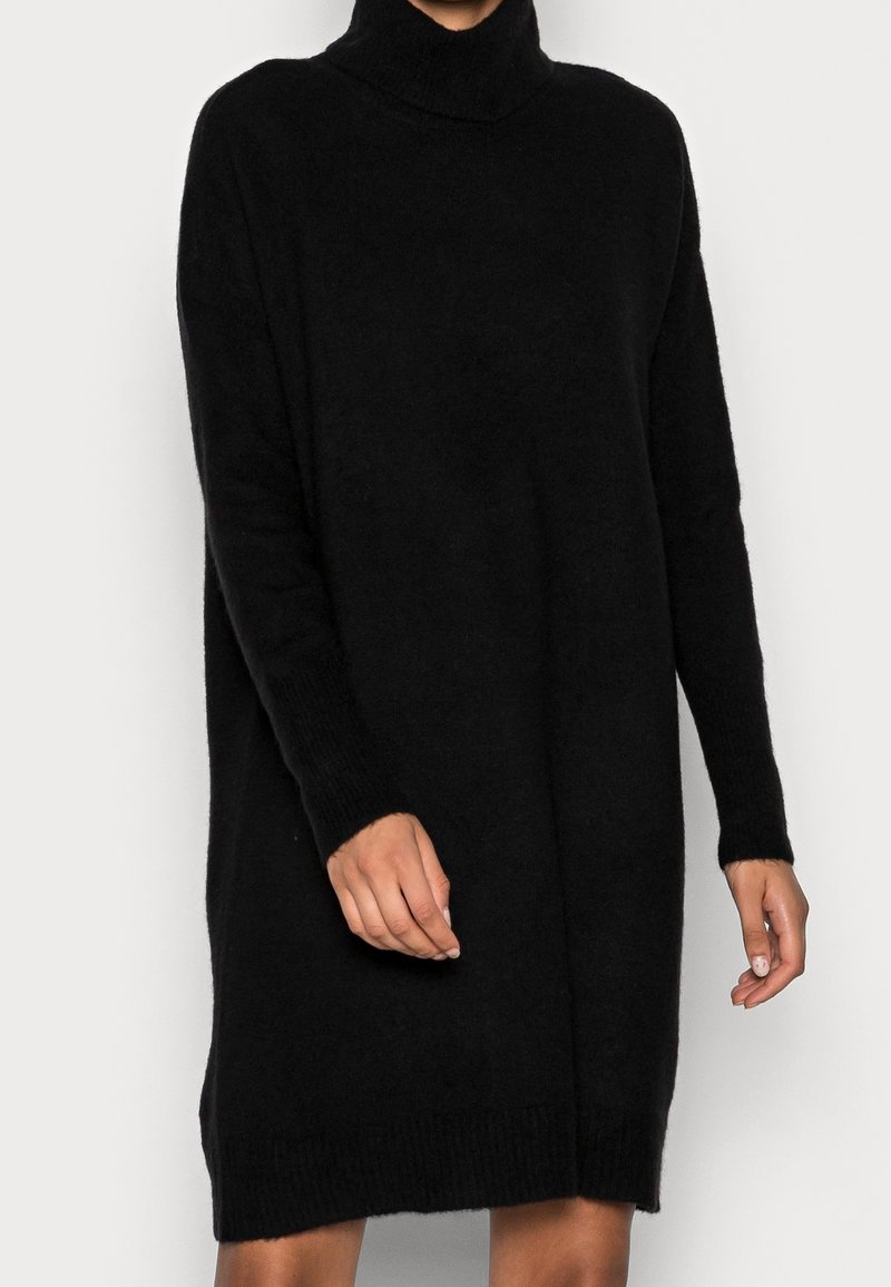 Femme portant une longue robe-pull noire à col roulé avec des manches longues, debout devant un fond clair uni.