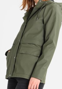 Oliven grøn vandtæt jakke med hætte, der har lynlås og trykknapslukning, to forlommer og en glat, struktureret finish.