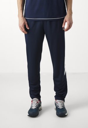 Marine joggerbroeken van gladde stof met een tapered fit en witte zijaccenten, gecombineerd met grijze en rode sportschoenen.