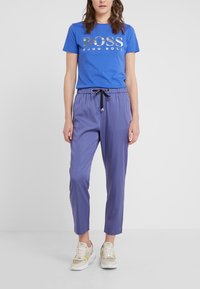 Niebieska koszulka z srebrnym logo "BOSS", w połączeniu z fioletowymi spodniami typu tapered z czarnym ściągaczem. Model nosi beżowe trampki.
