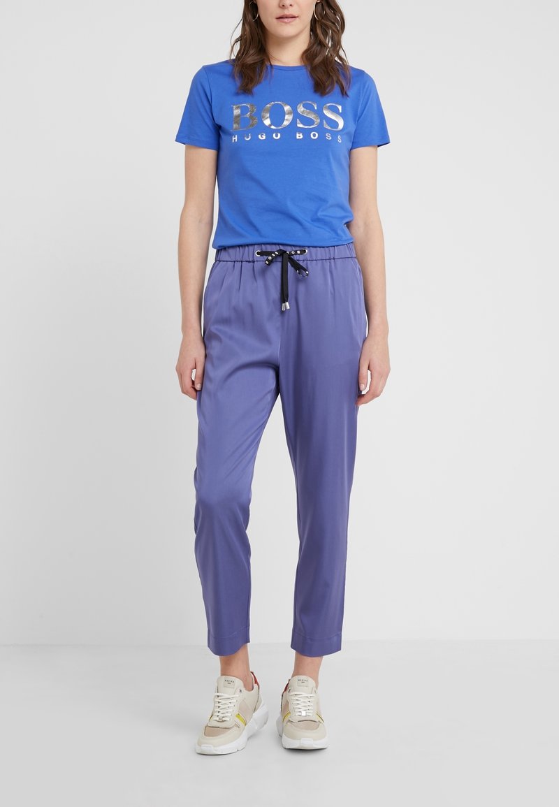 Niebieska koszulka z srebrnym logo "BOSS", w połączeniu z fioletowymi spodniami typu tapered z czarnym ściągaczem. Model nosi beżowe trampki.