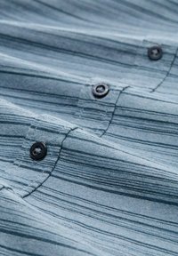 Textuurstof in tinten blauw met horizontale strepen. Bevat zwarte knopen en subtiele stiksels. Zachte, geweven stof.