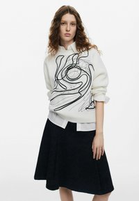 Suéter blanco de punto con bordado abstracto negro, superpuesto a una camisa blanca con cuello. Acompañado de una falda plisada negra con un patrón texturizado.