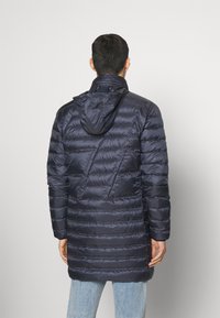 Mörkblå puffermantel med huva, med ett vadderat mönster, långa ärmar och en mellanhög längd, tillverkad av slät, glänsande tyg.