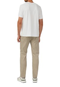 Man in een wit T-shirt met korte mouwen, beige broek en witte sneakers, staand met zijn rug naar de camera tegen een effen achtergrond.