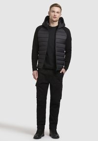 Gilet imbottito nero con cappuccio indossato sopra una maglietta a maniche lunghe nera, abbinato a pantaloni cargo neri e scarpe da ginnastica nere. Texture liscia e design elegante.