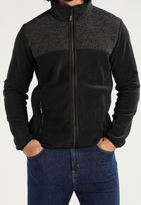 Schwarze Reißverschlussjacke mit strukturiertem grauem Einsatz oben, zwei Seitentaschen und hohem Kragen. Weicher Stoff und körpernahe Passform.