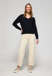 Schwarzer Baumwoll-V-Ausschnitt-Pullover mit langen Ärmeln, kombiniert mit cremefarbenen weit geschnittenen Hosen und beigen Turnschuhen. Verfügt über ein kleines rotes Logo-Accente.
