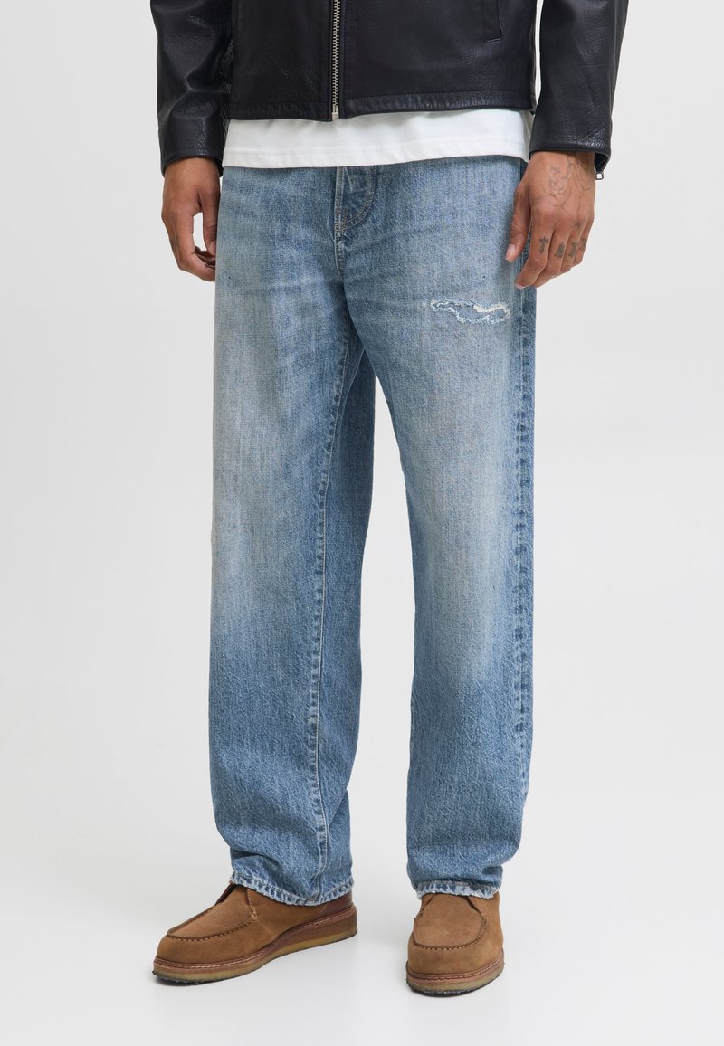 Mann trägt hellblaue, abgenutzte Jeans mit geradem Bein, braune Schuhe, ein weißes Hemd und eine schwarze Lederjacke mit sichtbaren Tattoos an den Händen.