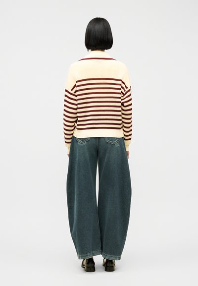 Personne aux cheveux courts noirs portant un pull rayé crème et bordeaux, un jean large bleu et des chaussures noires, vue de dos.