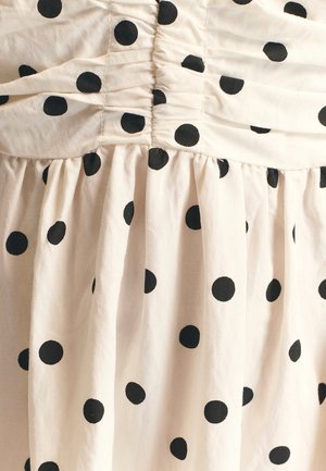 Kremfarvet bomuldskjole med en rynket overdel og en udvidet nederdel, prydet med store sorte polkaprikker over det hele. Blød tekstur.