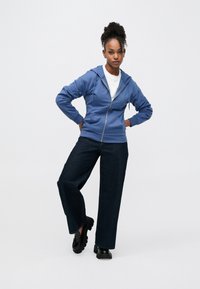 Μπλε φούτερ με φερμουάρ και χαλαρή εφαρμογή, συνδυασμένο με σκούρα τζιν φαρδύ πόδι και χοντρά μαύρα παπούτσια. Απαλή υφή και λείο ύφασμα.