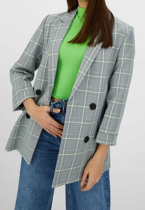 Vrouw draagt een grijs geruit colbert over een felgroene geribde top en blauwe spijkerbroek, met één hand in de taille gestoken.