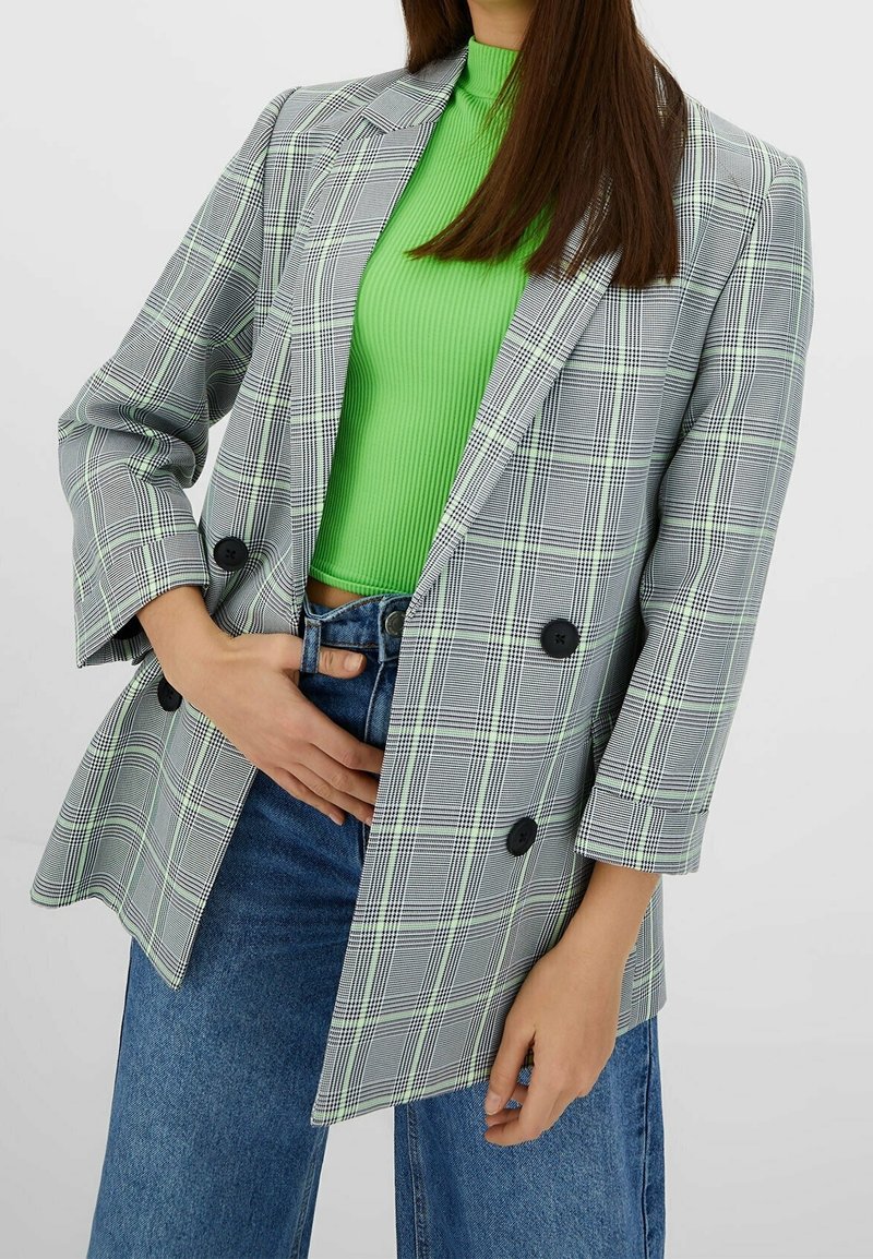 Femme portant un blazer à carreaux gris sur un top côtelé vert vif et un jean bleu, une main glissée à la taille.