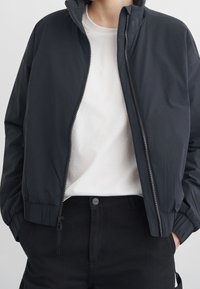 Personne portant une veste noire zippée par-dessus un t-shirt blanc et un pantalon noir, les mains dans les poches, montrée des épaules aux hanches.